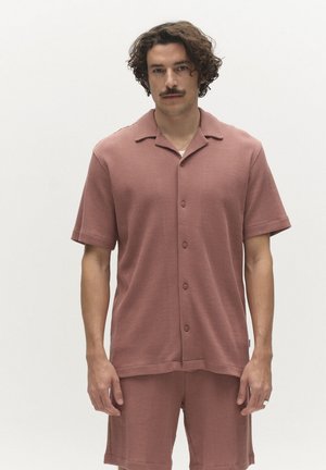 Moški z kodrastimi lasmi in brki, oblečen v ujemajočo se rožnato teksturirano srajco s kratkimi rokavi na gumbe in kratke hlače, stoječ ob enobarvni ozadju.