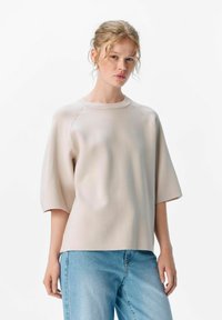 Lyse beige kortærmet top i et blødt, struktureret stof med rund halsudskæring og en afslappet pasform. Bæres med lyseblå jeans.
