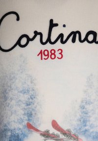 "Cortina 1983" texte au-dessus d'arbres bleus enneigés et de skis rouges sur un tissu tricoté blanc au thème des sports d'hiver.