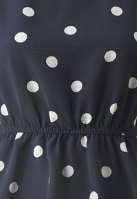 Tissu marine avec des pois blancs texturés. La taille froncée crée un effet blouson, mettant en valeur le tomber doux du tissu.