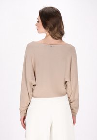 Top in maglia beige con scollo ampio e maniche lunghe, abbinato a pantaloni bianchi; texture liscia con un drappeggio casual.