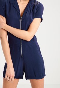 Marineblauer Kurzarm-Overall mit einem Frontreißverschluss, Kragen und umgeschlagenen Ärmeln. Hergestellt aus einem glatten Stoff mit lockerer Passform und vielseitigem Design.