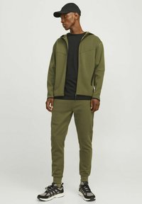 Survêtement de sport vert olive avec veste zippée, t-shirt noir et pantalons fuselés. Comprend des accents noirs et est accompagné de baskets noires.