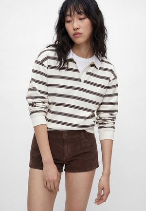 Młoda kobieta w białej i brązowej koszuli polo w paski z długim rękawem oraz brązowych sztruksowych szortach na jednolitym tle.