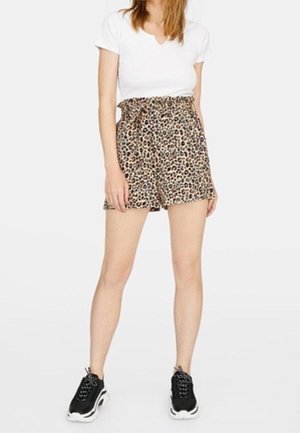 Kobieta ubrana w białą koszulkę z krótkim rękawem, wysokim stanem szorty w panterkę i czarne trampki stoi na białym tle.