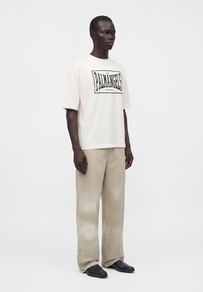 Άνδρας που φοράει μια oversized off-white μπλούζα με μαύρο κείμενο, μπεζ παντελόνια με φαρδιά μπατζάκια και κουμπιά κοντά στο τελείωμα, και μαύρα μυτερά παπούτσια.