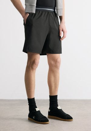 Oseba, oblečena v črne kratke hlače, vidno črno spodnje perilo z napisom Calvin Klein na pasu, črne nogavice in črne superge z gumijastimi podplati, stoji na sivem tleh.