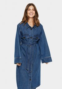 Denim dlhý kabát s opaskom v páse, veľkými vreckami a gombíkovým zapínaním. Má golier a široké rukávy v stredne modrej farbe.