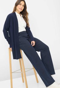 Marineblå cardigan i blødt strikket materiale med en lang, åben front, parret med højtaljede marineblå bukser og lyse sneakers, siddende på en skammel.