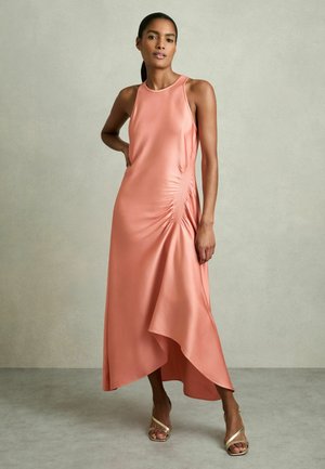 Mujer de pie con un vestido midi coral sin mangas con detalle fruncido a los lados y sandalias doradas de tacón con tiras, sobre un suelo gris.