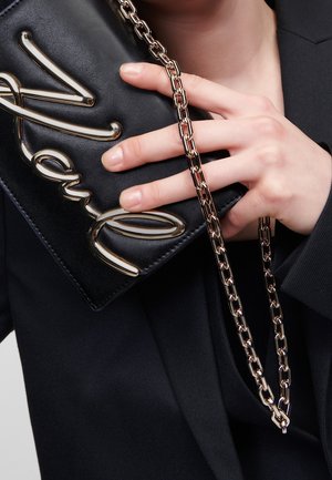 Mano sosteniendo una bolsa de cuero negro con una correa de cadena dorada y grandes letras doradas en cursiva en la solapa de la bolsa.