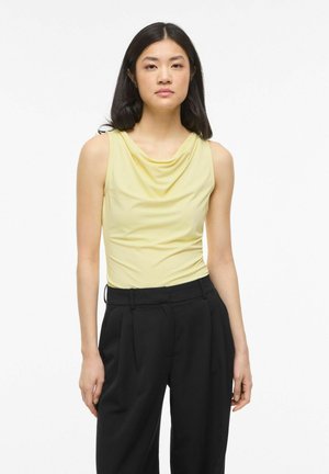 Kobieta o prostych czarnych włosach, ubrana w bezrękawnikowy blady żółty drapowany top i czarne plisowane spodnie z wysokim stanem, stojąca na jednolitym białym tle.