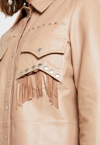 Beige läderjacka med krage, tryckknappar, två bröstfickor, silvernitar och fransdetaljer på fickkanterna.