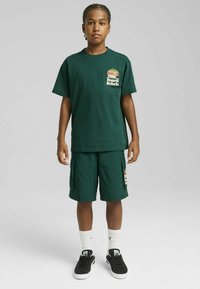 Camiseta de algodón verde con un pequeño logo, combinada con pantalones cortos de cargo a juego. Zapatillas negras y calcetines blancos completan el conjunto.