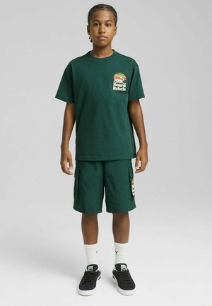Camiseta de algodón verde con un pequeño logo, combinada con pantalones cortos de cargo a juego. Zapatillas negras y calcetines blancos completan el conjunto.