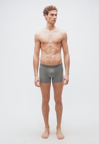 Νεαρός άνδρας που στέκεται ξυπόλυτος φορώντας γκρι μποξεράκια, κοιτάζοντας προς τα πλάγια πάνω σε απλό λευκό φόντο.