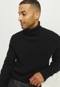 Schwarzer Rollkragenpullover mit gerippter Textur, langen Ärmeln und einem entspannten Schnitt, getragen von einer Person vor einem neutralen Hintergrund.