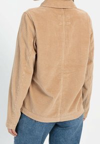 Veste en velours côtelé beige avec une surface texturée à côtes, col rabattu et manches longues, présentant un ourlet droit et un détail de couture au dos.