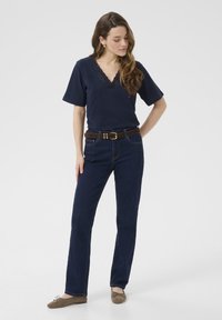 Marineblå kortærmet bluse med blonder ved V-halsen, parret med lige skårne mørkeblå jeans og et brunt læderbælte; modellen står med den ene hånd på hoften.