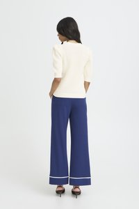 Femme aux longs cheveux noirs portant un haut en maille crème avec des manches bouffantes et un pantalon large bleu marine avec un ourlet blanc, debout les mains dans les poches.