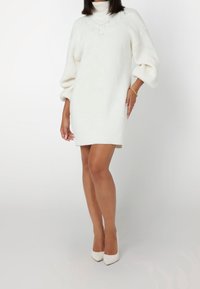 Abito maglione bianco con collo alto, motivo testurizzato sul petto e maniche a palloncino, abbinato a décolleté bianchi a punta.