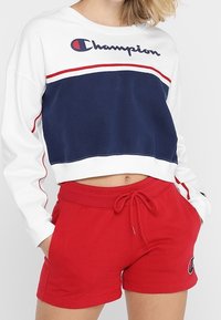 Sudadera blanca corta con franja horizontal azul marino y acentos rojos; presenta el logo de "Champion", combinada con pantalones cortos rojos con detalle del logo.