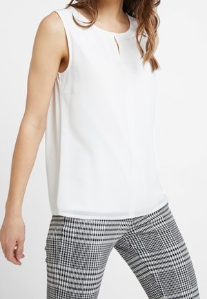 Kobieta w białej, bezrękawnikowej bluzce z małym przednim rozcięciem i czarno-białych, w kratę spodniach na białym tle.