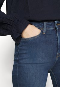 Dunkelblaue Denim-Jeans mit schmaler Passform, ausgestattet mit einer Fronttasche, dezenten Nähten und einer strukturierten schwarzen Bluse mit langen Ärmeln.