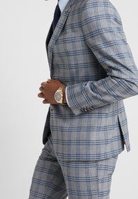 Completo a quadri grigio e blu con vestibilità su misura, caratterizzato da un giubbotto a un bottone e pantaloni abbinati. Accentuato da un orologio d'oro al polso.