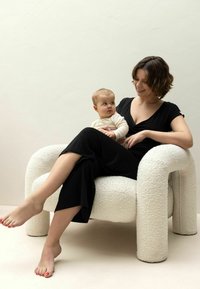 Fauteuil lounge blanc texturé avec des bras et des jambes arrondis, occupé par une femme en tenue noire tenant un bébé de couleur claire.