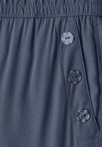 Close-up of a dark gray fabric waistband with gathered elastic and a vertical row of three large glossy two-hole buttons on a pocket flap.