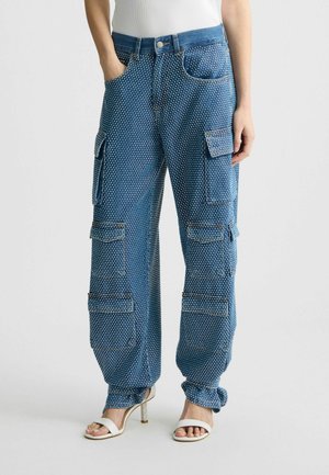 Personne portant un pantalon cargo bleu à pois avec plusieurs poches et des sandales blanches à talons, debout devant un fond uni.