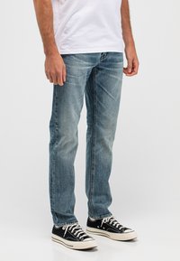 Mann trägt hellblaue, ausgewaschene Jeans und schwarze Low-Top-Sneaker mit weißen Schnürsenkeln, steht vor einem schlichten weißen Hintergrund.