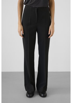 Osoba ubranа w czarne proste spodnie wizytowe i czarne buty, stojąca na szarej podłodze na tle jednolitego, jasnego tła.