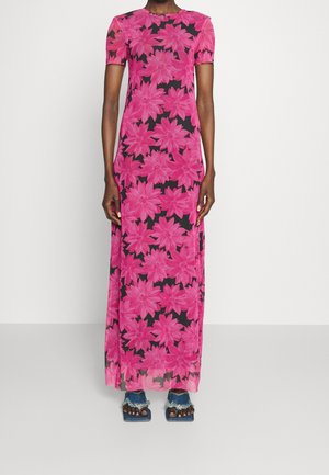 Osoba ubrana w czarną sukienkę z krótkim rękawem, sięgającą do ziemi, z dużym różowym wzorem kwiatowym oraz niebieskie, postrzępione sandały na odkryte palce.