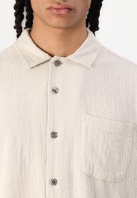 Light beige button-up shirt with a textured fabric, collar, and single chest pocket. Features gray buttons and a relaxed fit design.