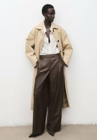 Beige oversized trenchcoat over een witte gestreepte blouse met een diepe V-hals, gecombineerd met donkerbruine leren wijde broek.