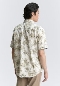 Chemise à manches courtes avec boutons, de couleur beige clair, ornée d'un imprimé de palmier en marron foncé et vert. Texture douce, coupe décontractée et ourlet arrondi.