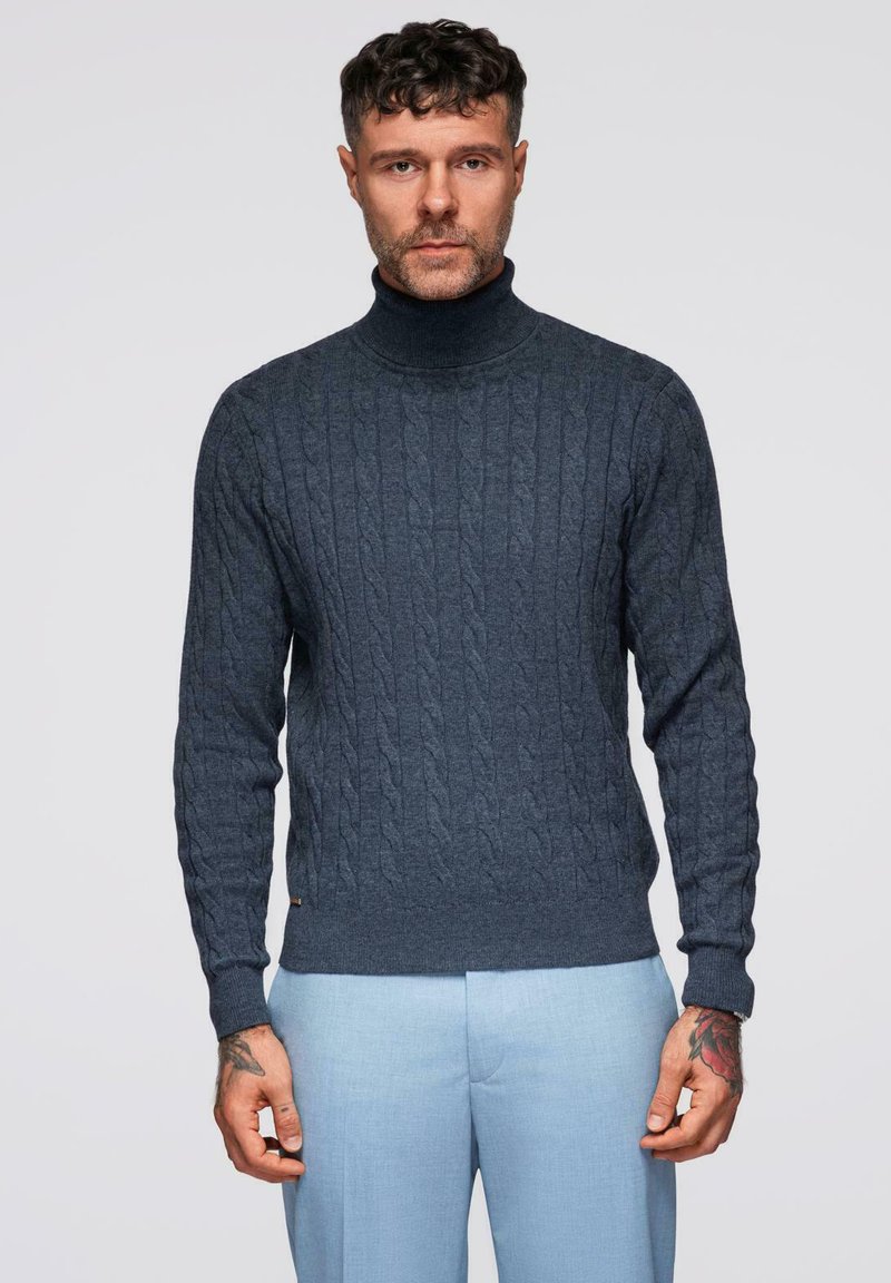 Pull à col roulé bleu marine en tricot texturé avec un motif en cable, associé à un pantalon bleu clair. Détails discrets de la marque au bas.