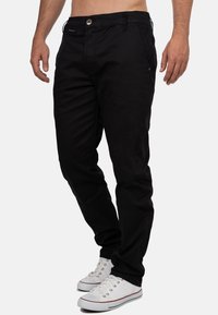 Pantaloni neri a gamba dritta con chiusura a bottone e cerniera, tasche frontali e tessuto in cotone morbido. Indossati con scarpe da tennis bianche.