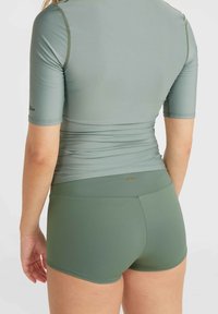 Rash guard vert à manches courtes et short de bain assorti, fabriqués en tissu lisse et extensible avec des coutures plates et un détail de marque subtil.