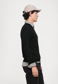 Schwarzer Strickpullover mit Diamantmuster, darüber ein kariertes Hemd. Eine beige Mütze mit verstellbarem Riemen rundet das Outfit ab.