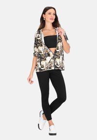 Sort og guld mønstret zip-up jakke med korte ærmer over en sort crop top, parret med sorte leggings og hvide sneakers.