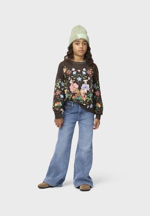 Fille portant un bonnet en laine vert, un sweat-shirt sombre à motifs floraux avec des dessins de chatons, un jean bleu à jambes larges et des chaussures marron, debout sur un fond gris.