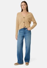 Beige gestrickte Strickjacke mit V-Ausschnitt, drei Knöpfen und gerippten Bündchen, kombiniert mit hellblauen weit geschnittenen Jeans und beige-farbenen Slippers.
