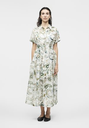 Γυναίκα που στέκεται φορώντας φόρεμα με ζώνη, κοντά μανίκια, κουμπιά και στροβιλιστό αφηρημένο σχέδιο σε πράσινο, χρυσό και μπλε, καθώς και μαύρα επίπεδα παπούτσια.