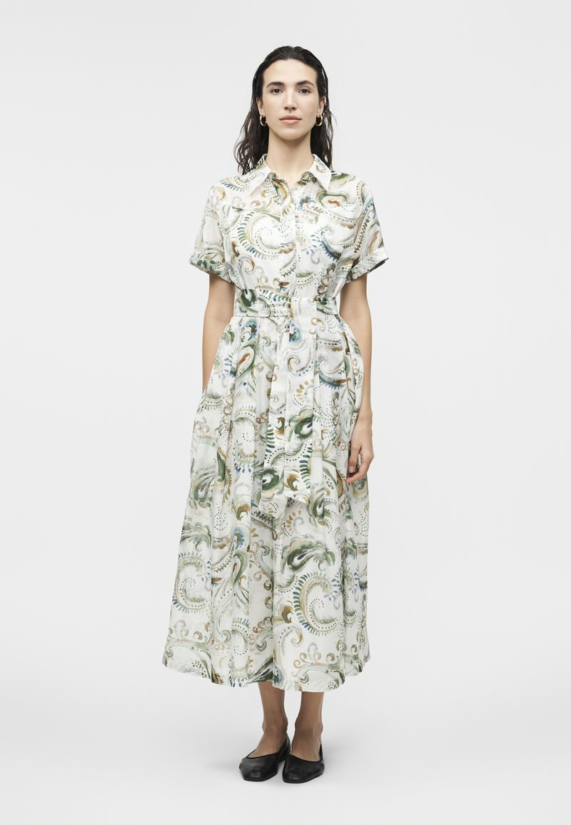 Γυναίκα που στέκεται φορώντας φόρεμα με ζώνη, κοντά μανίκια, κουμπιά και στροβιλιστό αφηρημένο σχέδιο σε πράσινο, χρυσό και μπλε, καθώς και μαύρα επίπεδα παπούτσια.