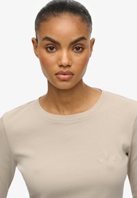 Haut à manches longues beige côtelé avec un col rond et un logo brodé discret sur la poitrine. Texture lisse, coupe ajustée.