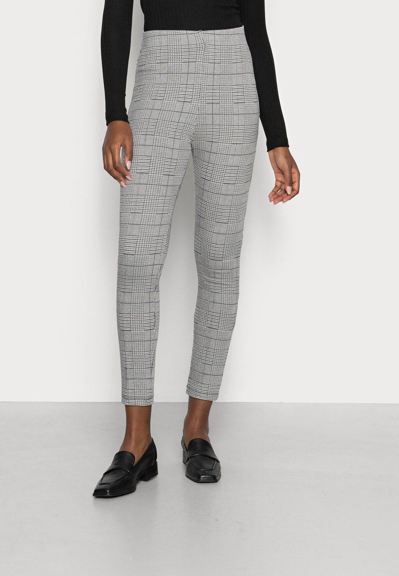 Personne portant un pantalon gris à carreaux ajusté taille haute, un haut noir à manches longues et des mocassins en cuir noir, debout devant un fond blanc uni.