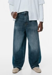 Široke traperice u izblijedjeloj tamnoplavoj boji s opuštenim krojem. Iznad ruba su izribane, a suptilno bora dodaju teksturu. Nošene s bež tenisicama.